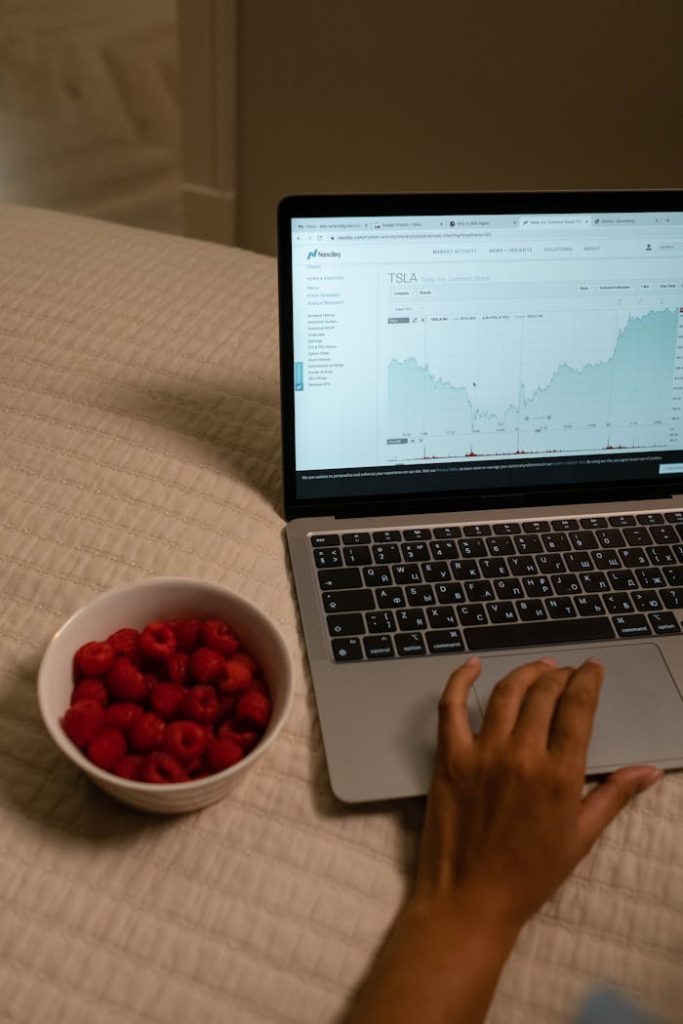 Laptop showing stock market data with hand using it and bowl of raspberries beside.