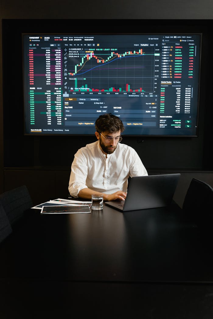 A focused man analyzing stock market trends on a laptop in an office setting with charts displayed.